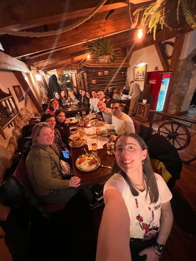 Un gruppo WeRoad sorride per un selfie mentre cena insieme a un lungo tavolo all'interno di un ristorante rustico e illuminato in modo accogliente.