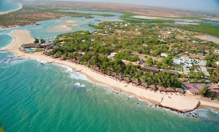 Eine Luftaufnahme einer tropischen Küste mit einem Sandstrand, türkisfarbenem Wasser und einem Resort, eingebettet zwischen grünen Bäumen und einer Lagune.