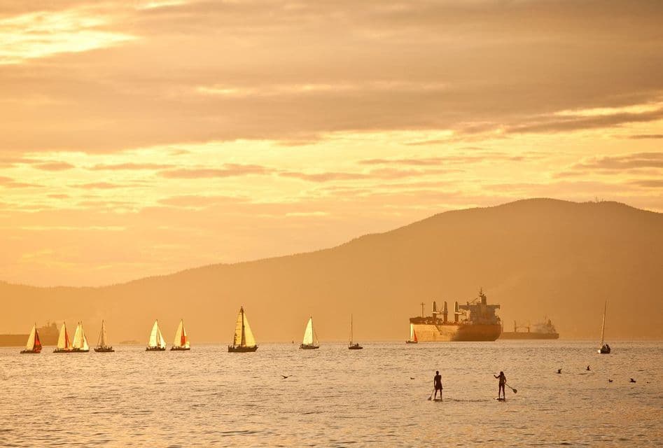 Two people stand-up paddleboarding on the water near sailboats and a large cargo ship during a golden sunset.