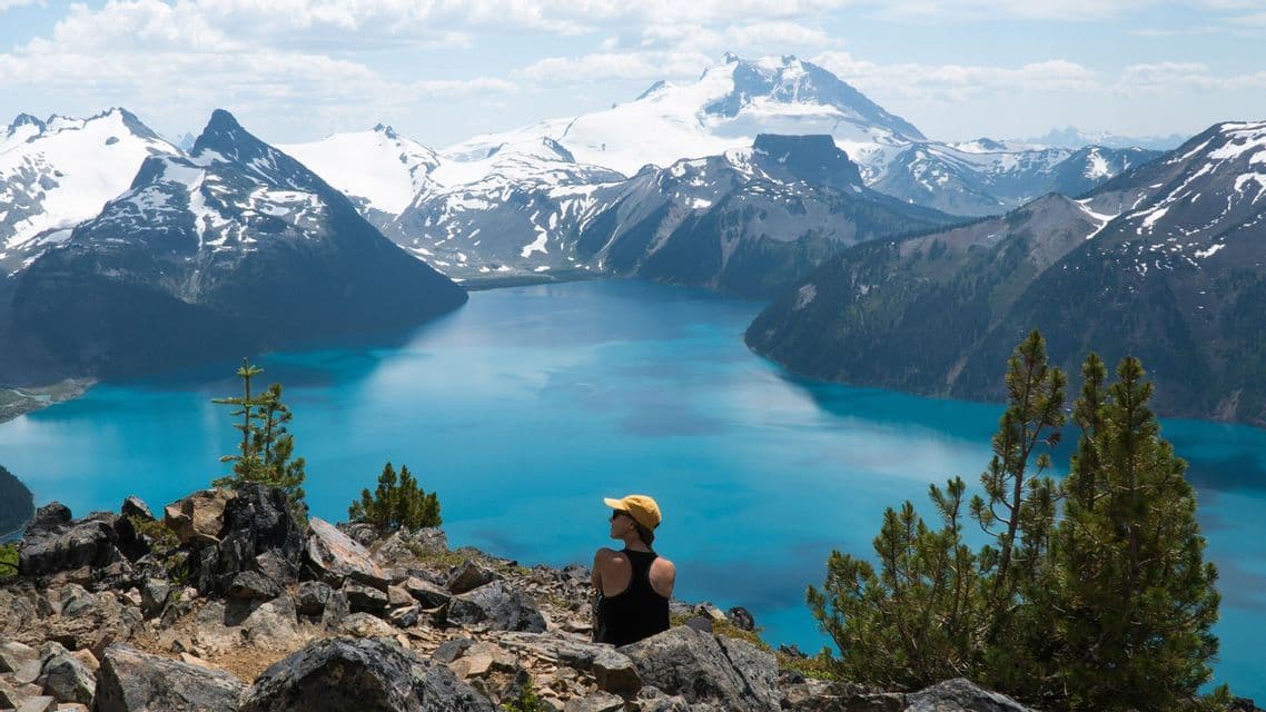 A person in a yellow cap sits on a rocky ledge, looking out over a turquoise alpine lake surrounded by snow-capped mountains.