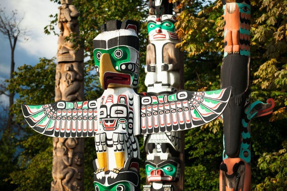 Colorful carved wooden totem poles, including one of an eagle with spread wings, stand among green trees.