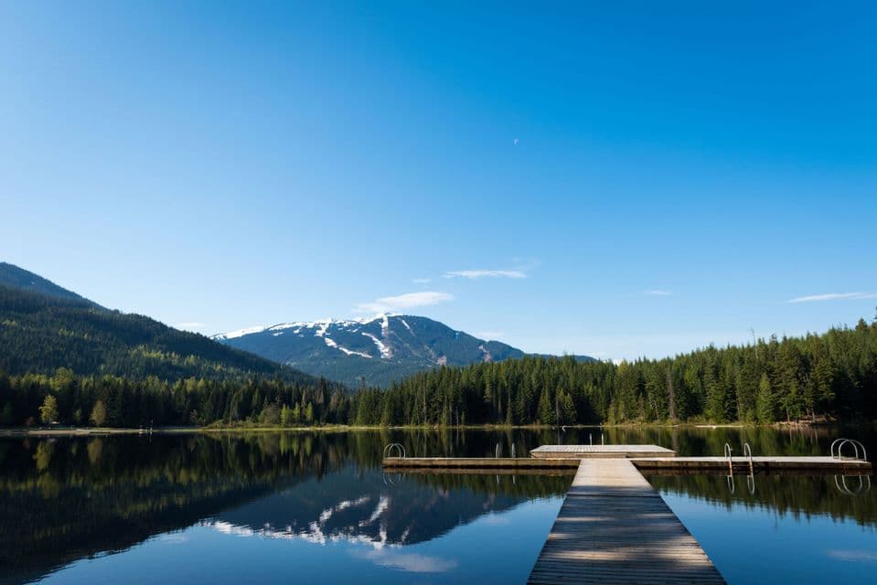 A wooden dock on a calm lake reflects a pine forest and a snow-capped mountain against a clear blue sky.
