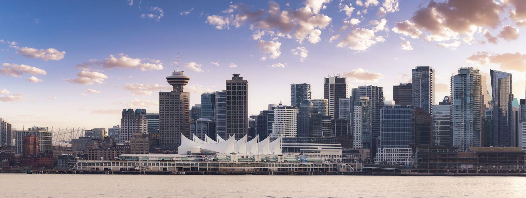 A panoramic view of a city skyline on a waterfront, with modern skyscrapers and a pier under a partly cloudy sky.