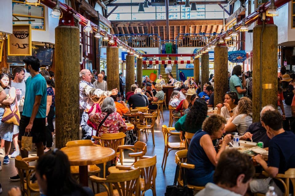 A crowd of people eating and socializing inside a bustling indoor food market with large wooden pillars.