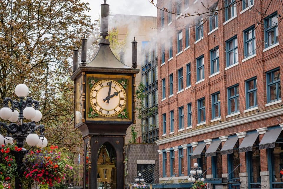 A vintage steam clock releases steam on a city street corner next to a red brick building and trees.