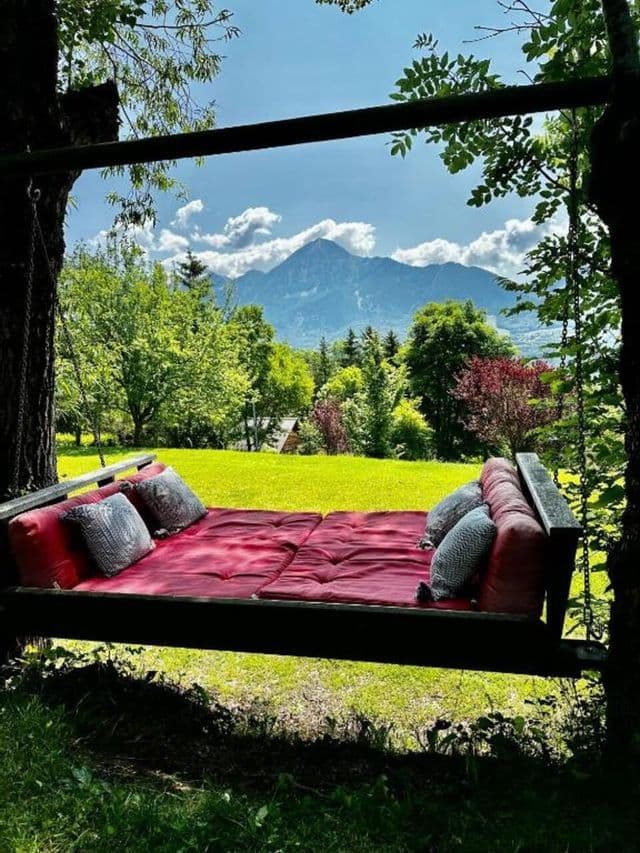 Un grand lit suspendu avec des coussins rouges surplombe une pelouse verte et une montagne lointaine sous un ciel partiellement nuageux.