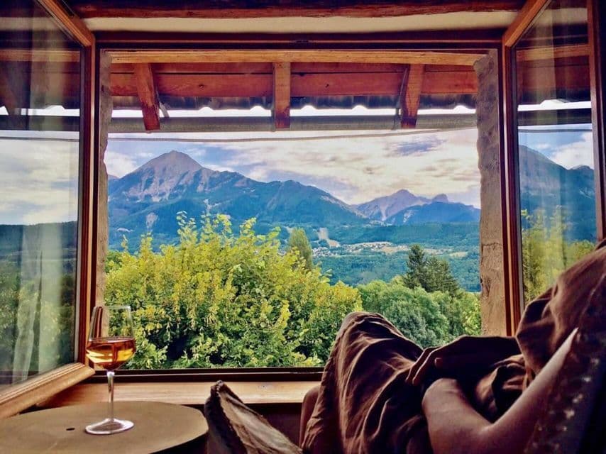 Une personne se détend avec un verre de vin près d'une fenêtre ouverte donnant sur une vallée montagneuse verdoyante.