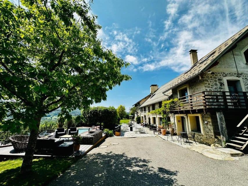 Un bâtiment rustique en pierre avec une cour pavée et une terrasse de piscine dotée de mobilier de détente, entouré d'arbres verdoyants sous un soleil éclatant.