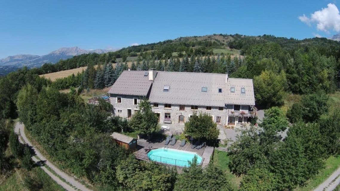 Une vue aérienne d'une grande maison en pierre avec une piscine, nichée parmi des arbres verts sur une colline avec des montagnes en arrière-plan.