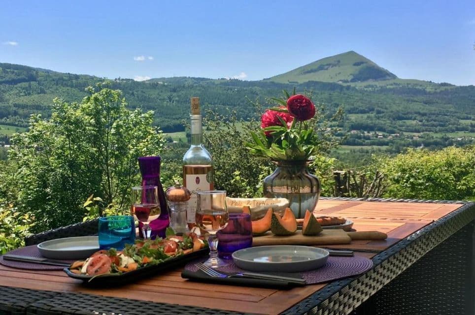 Une table en plein air est dressée avec des mets et du vin rosé, offrant une vue sur une vallée verdoyante et une grande montagne.