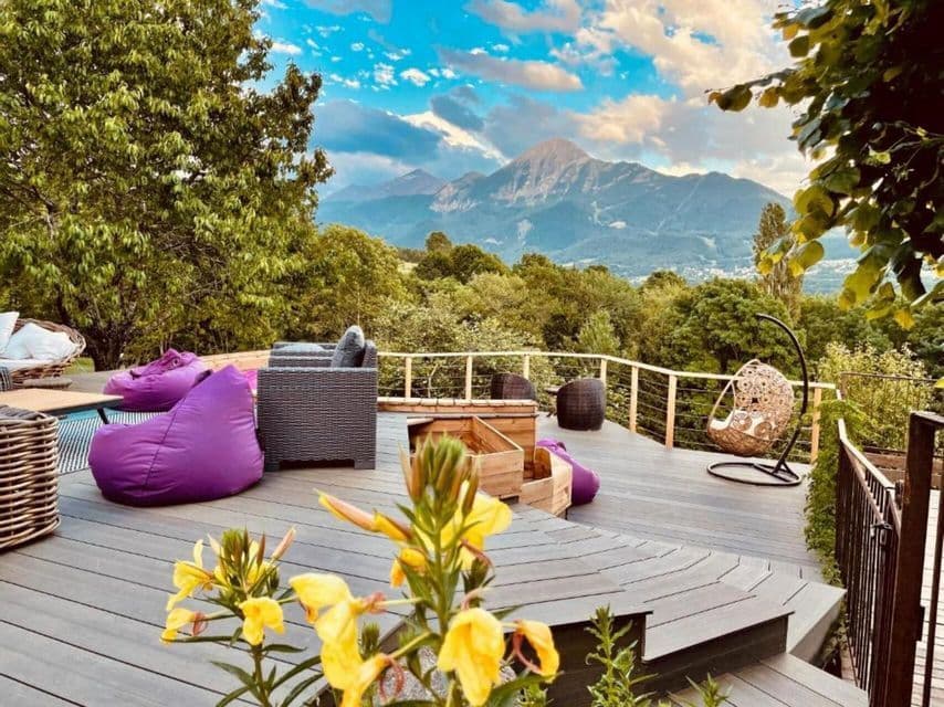 Une terrasse en bois avec des meubles de salon et des poufs violets surplombe une chaîne de montagnes boisée sous un ciel bleu.