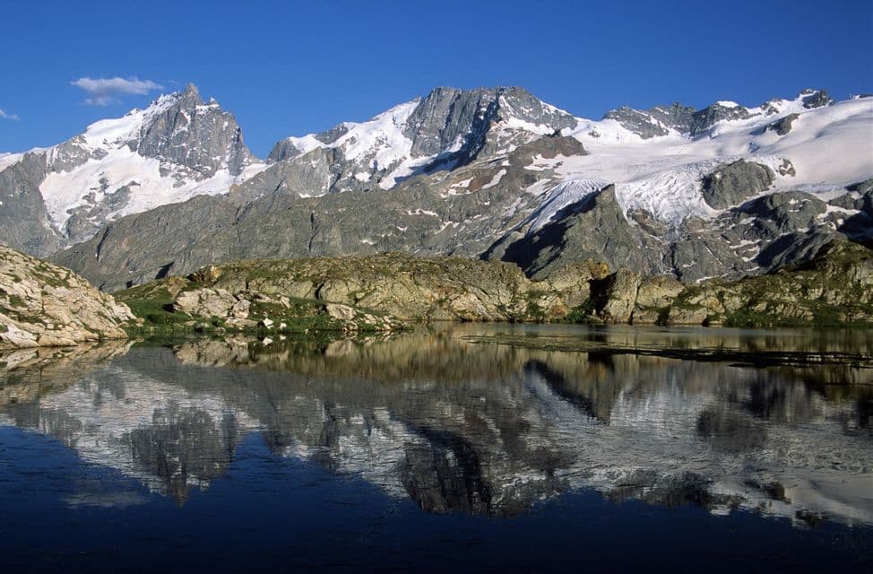 Une chaîne de montagnes enneigée se reflète parfaitement dans l'eau calme d'un lac alpin sous un ciel bleu clair.