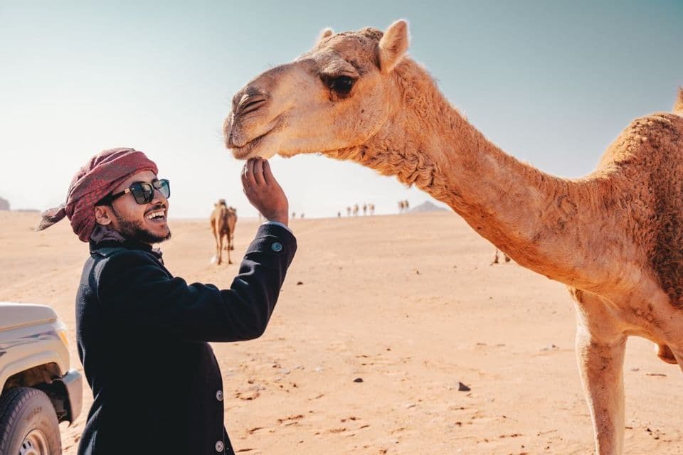 Ein lächelnder Mann mit rotem Turban und Sonnenbrille berührt sanft das Kinn eines Kamels in einer sonnigen Wüstenlandschaft.