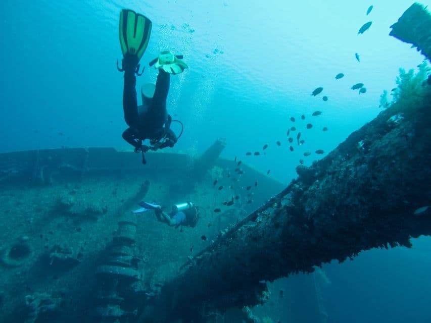 Two divers from a WeRoad group trip explore a sunken shipwreck underwater, with schools of small fish swimming by.
