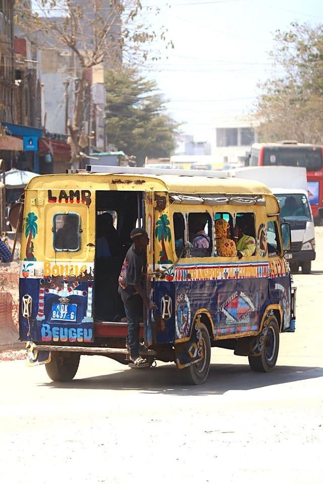 Ein Mann hängt sich an das Heck eines bunt bemalten Minibusses, der mit Fahrgästen eine Stadtstraße entlangfährt.
