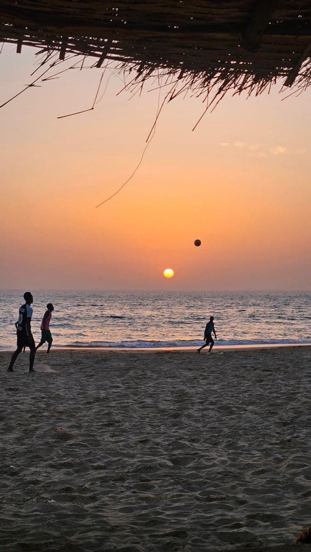 Silhouetten von Fußball spielenden Menschen an einem Sandstrand bei Sonnenuntergang, mit dem Ball in der Luft über dem Meer.