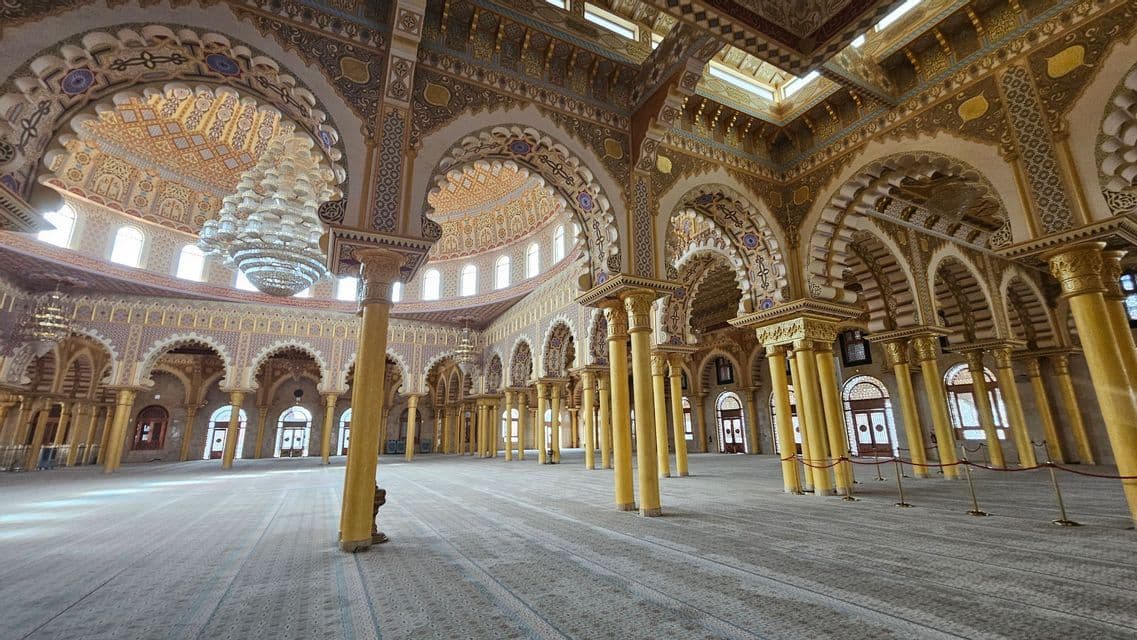 Der weitläufige Innenraum einer Moschee mit Reihen goldener Säulen, verzierten Bögen und großen Kronleuchtern über einem gemusterten Teppich.