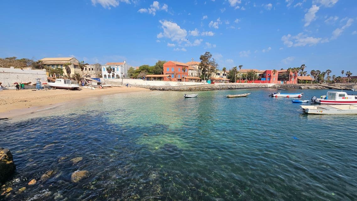 Kleine Boote liegen in einer ruhigen Bucht mit klarem, türkisfarbenem Wasser, einem Sandstrand und farbenfrohen Gebäuden am Ufer.
