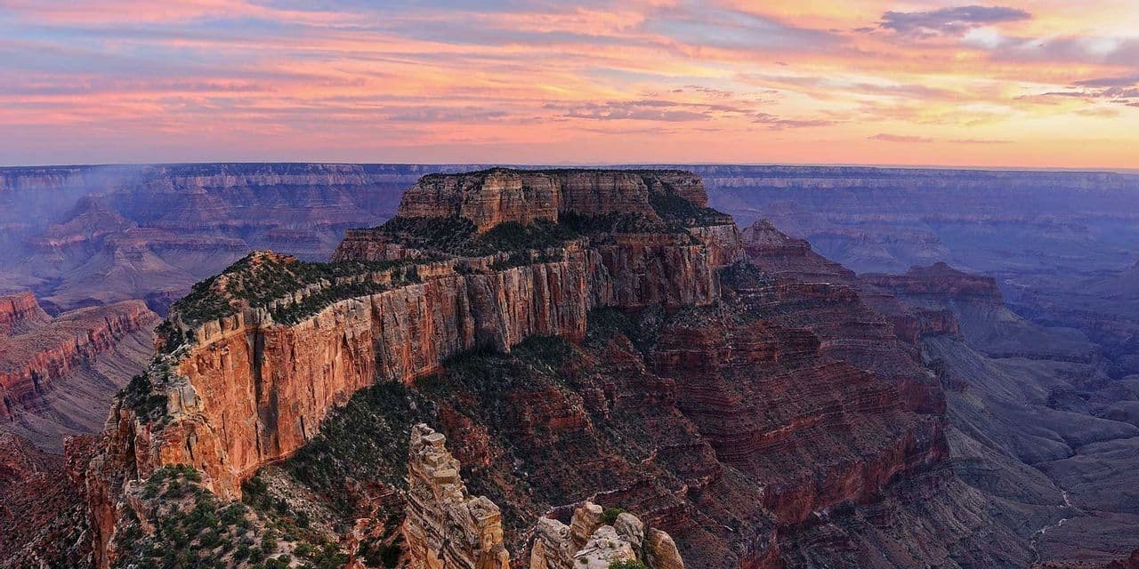 A panoramic view of a vast canyon with layered rock formations under a colorful pink and orange sunset sky.
