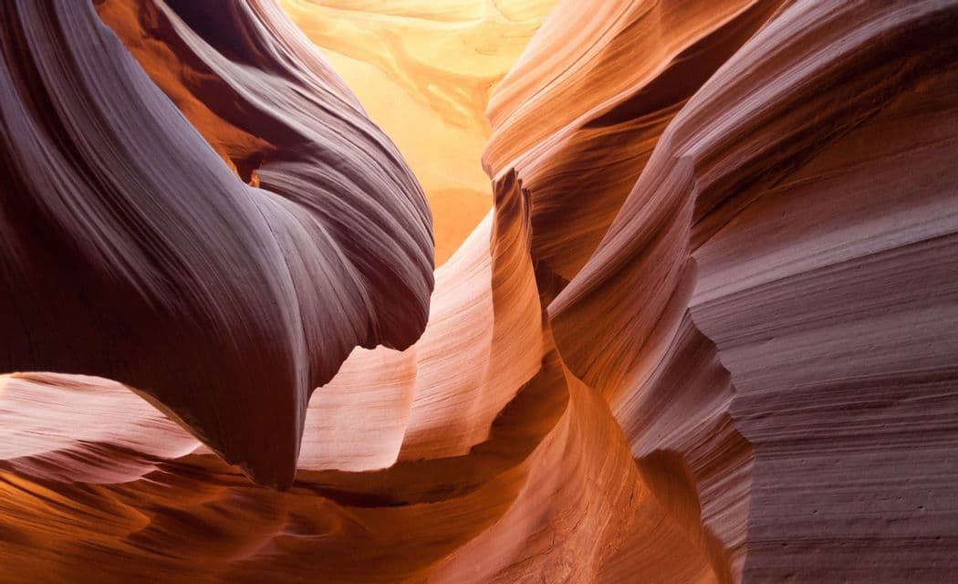 Sunlight filters into a narrow slot canyon, illuminating the smooth, layered sandstone walls in tones of red and orange.