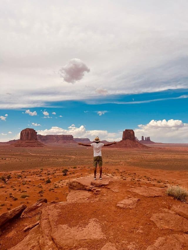 A person in a hat stands with arms outstretched on a rocky ledge, overlooking a vast desert landscape with large rock formations.