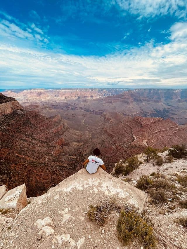 A person wearing a WeRoad t-shirt sits on a rocky overlook, admiring the view of a vast canyon under a blue and cloudy sky.