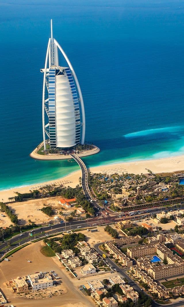 Luftaufnahme eines großen, segelförmigen Gebäudes auf einer künstlichen Insel, das über eine Brücke mit einem sandigen Küstenstreifen verbunden ist.