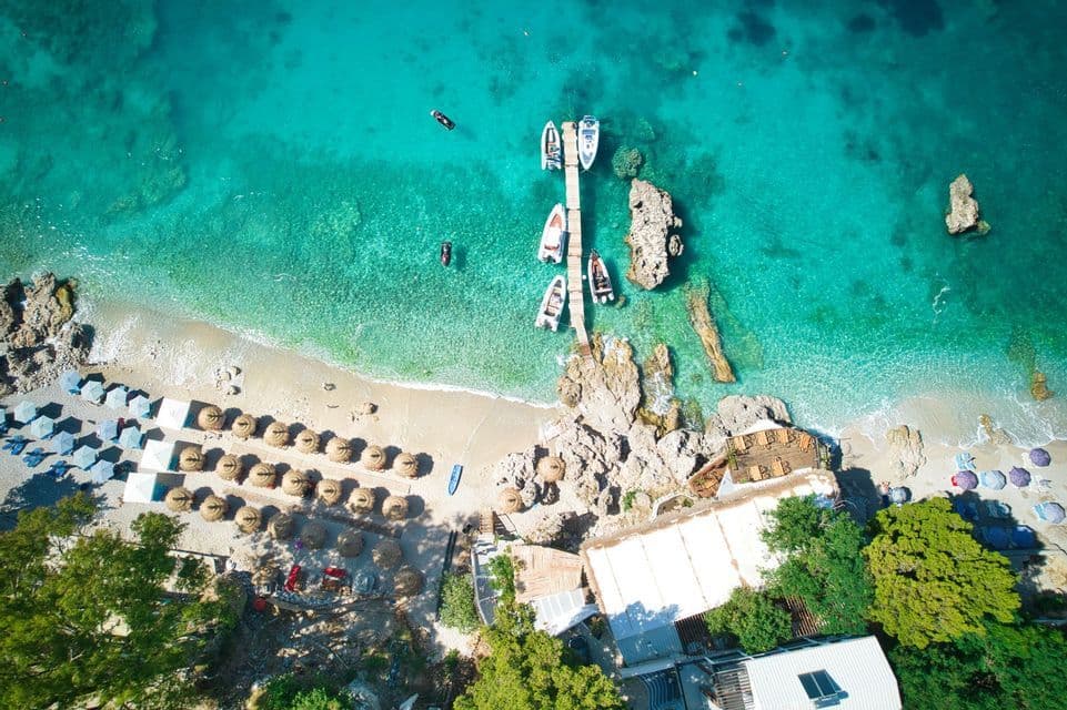 Luftaufnahme eines sonnigen Strandes mit klarem türkisen Wasser, einem Holzsteg mit Booten und Reihen von Strohschirmen.