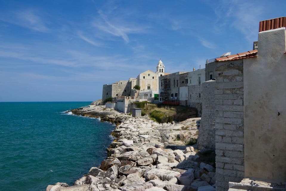 Una città di mare con edifici in pietra e una chiesa costruita su una costa rocciosa accanto al mare turchese sotto un cielo blu.
