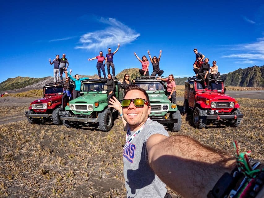 Un homme prend un selfie avec un groupe WeRoad posant sur et autour de jeeps tout-terrain colorées dans un vaste paysage aride sous un ciel bleu.