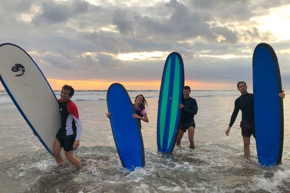 Eine WeRoad-Gruppe von vier Personen mit Surfbrettern steht bei Sonnenuntergang im Meer.