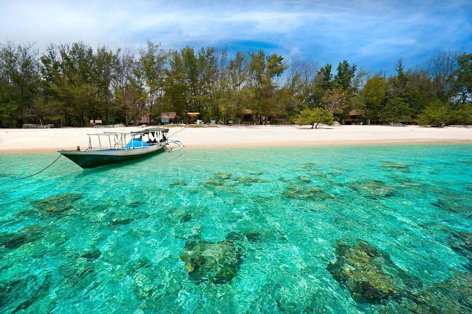 Un bateau est mouillé dans des eaux peu profondes, claires et turquoise, au large d'une plage de sable blanc bordée d'arbres verts luxuriants, sous un ciel bleu.