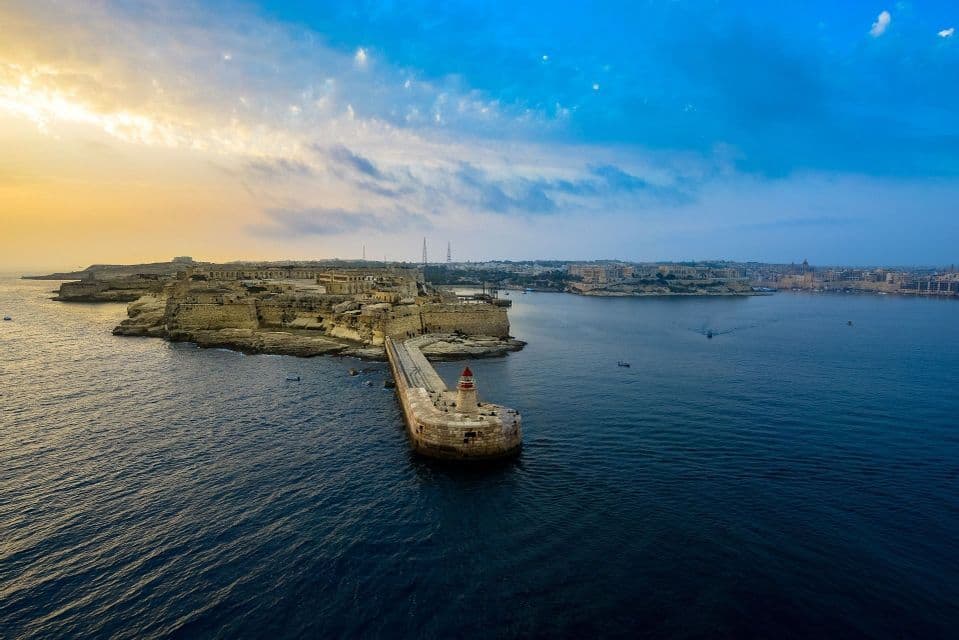Luftaufnahme eines steinernen Wellenbrechers und Leuchtturms, die sich von einer befestigten Küstenstadt bei Sonnenuntergang ins Meer erstrecken.