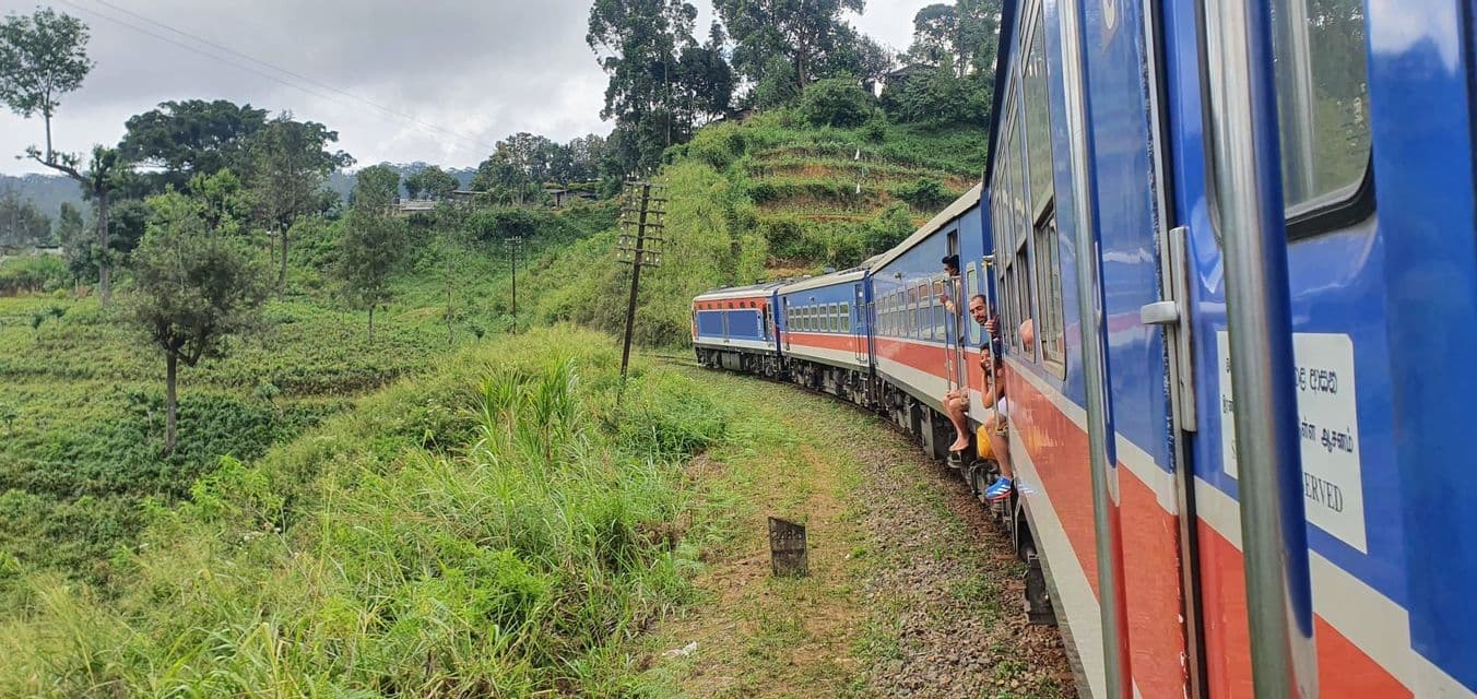 A WeRoad group trip sits in the open doorways of a blue and red train traveling through a lush, green, hilly landscape.