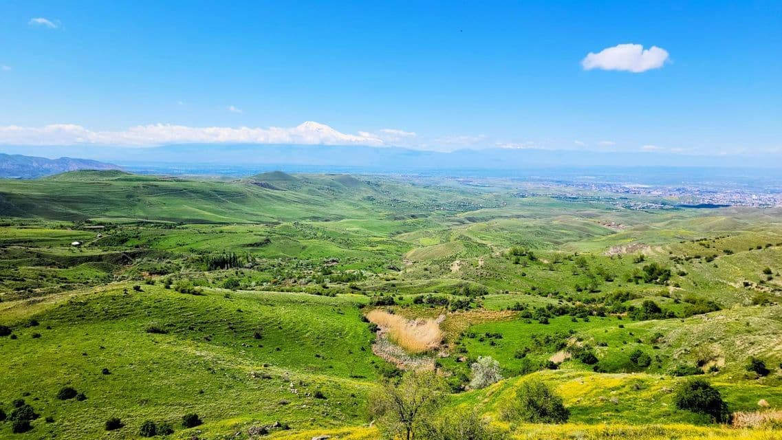 Une vue panoramique de collines verdoyantes s'étendant vers une montagne enneigée lointaine sous un ciel bleu éclatant.