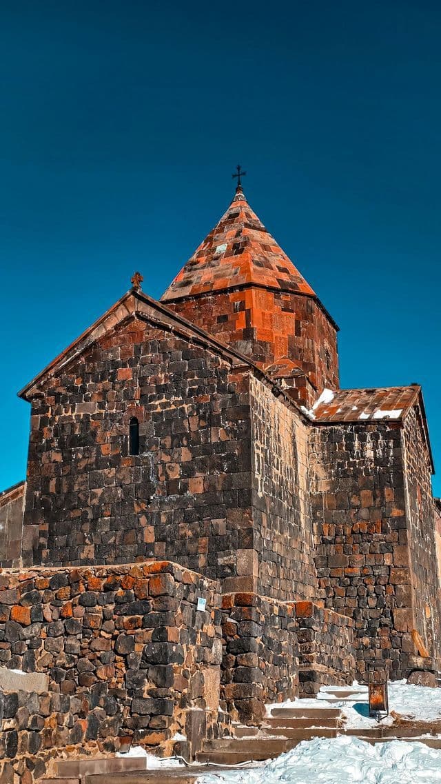 Un monastère historique en pierre au toit conique se dresse sous un ciel d'un bleu profond, avec de la neige recouvrant le sol alentour.