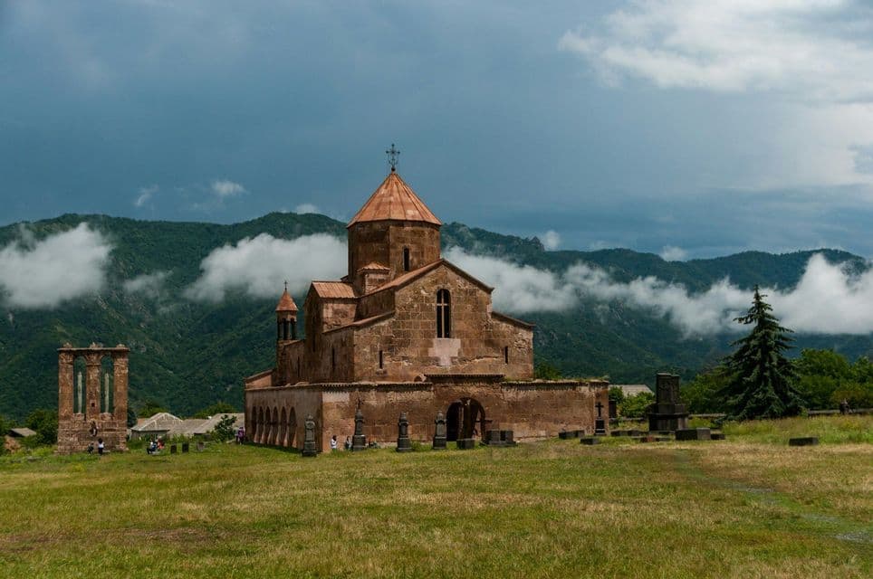 Un ancien monastère en pierre avec un toit en dôme se dresse dans un champ herbeux, avec des collines verdoyantes et des nuages bas en arrière-plan.