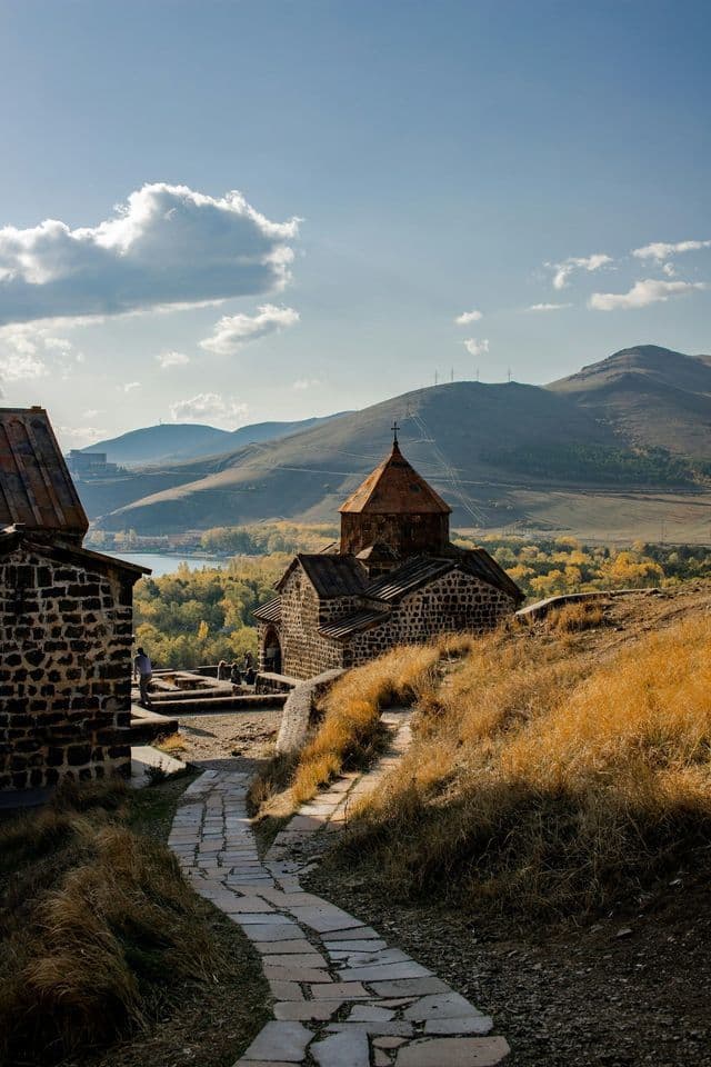 Un chemin pavé sur une colline herbeuse mène à deux anciennes églises en pierre avec des montagnes en arrière-plan.