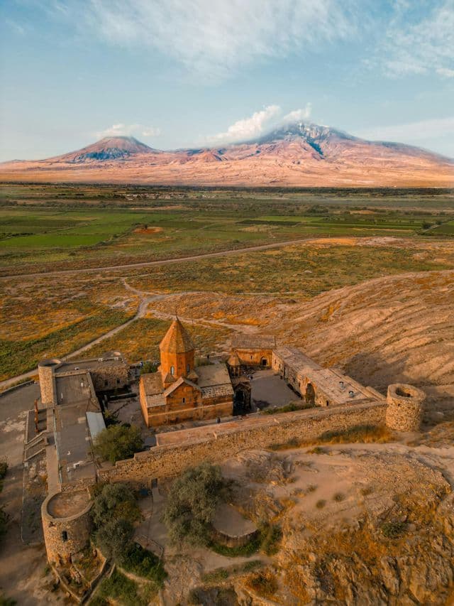 Vue aérienne d'un monastère historique en pierre sur une colline, dominant un vaste paysage avec une grande montagne à double sommet en arrière-plan.