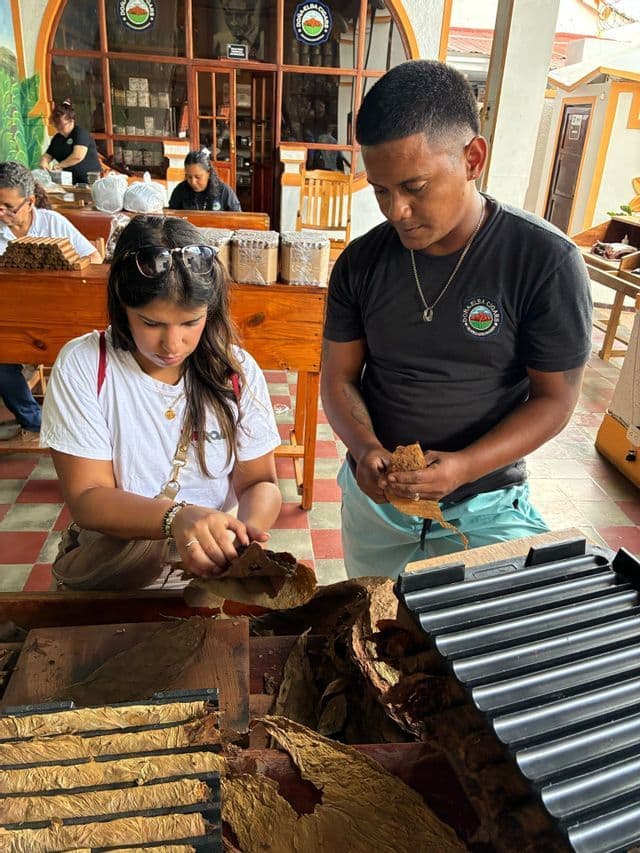 Une femme lors d'un voyage de groupe WeRoad apprend à rouler un cigare à la main auprès d'un homme dans un atelier avec des feuilles de tabac séchées.