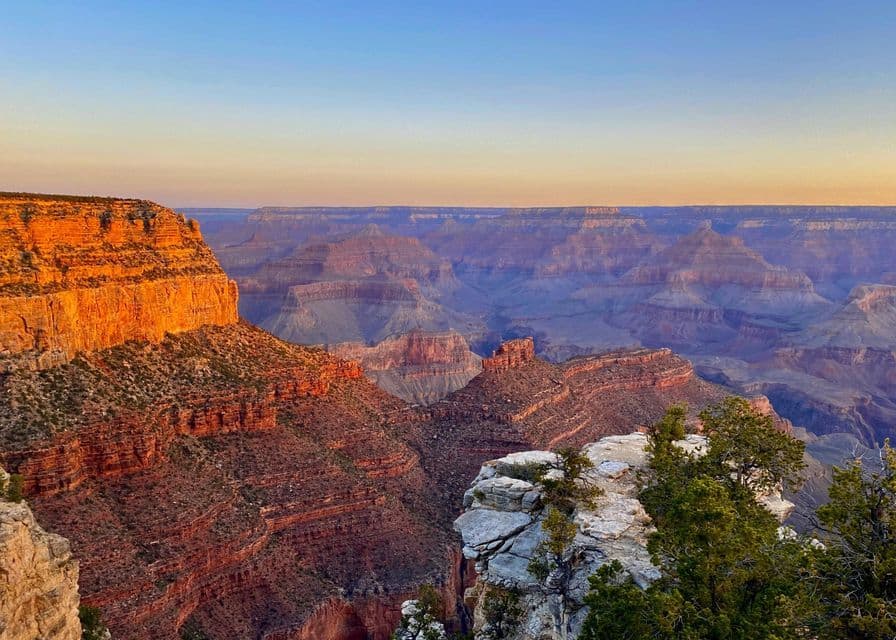 A vast, layered canyon with rock formations lit by warm sunrise light under a clear sky.