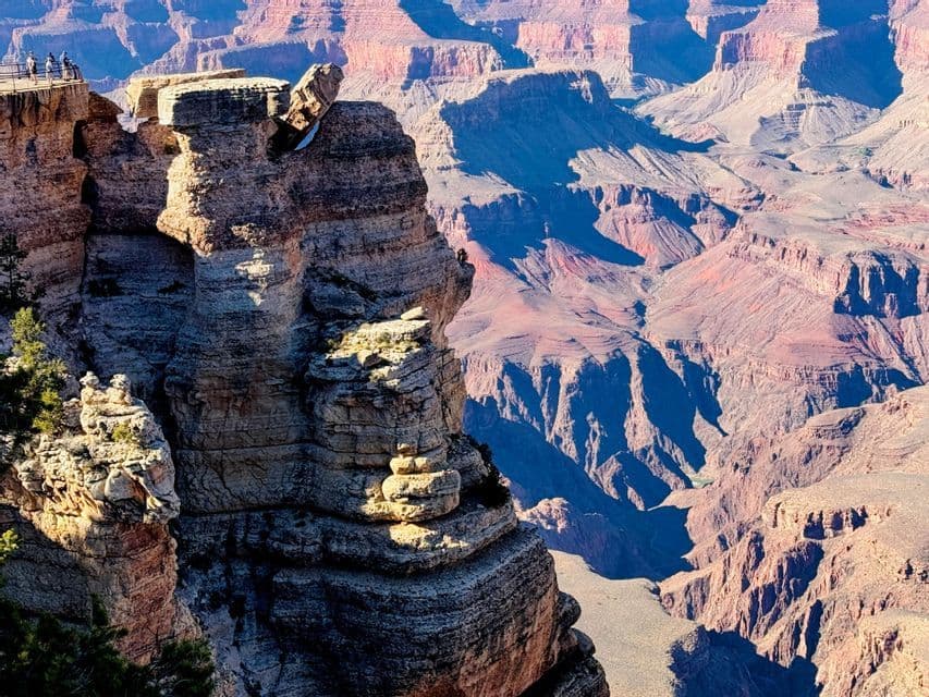 A vast canyon with sunlit, layered red rock formations, featuring a large balanced rock and a distant viewing platform with people.