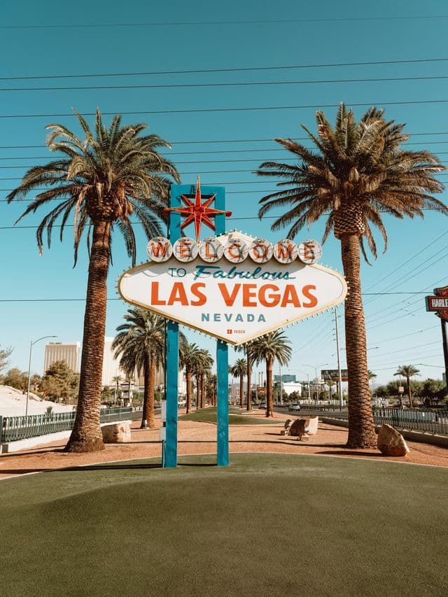 The 'Welcome to Fabulous Las Vegas' sign is framed by two tall palm trees against a clear blue sky on a sunny day.