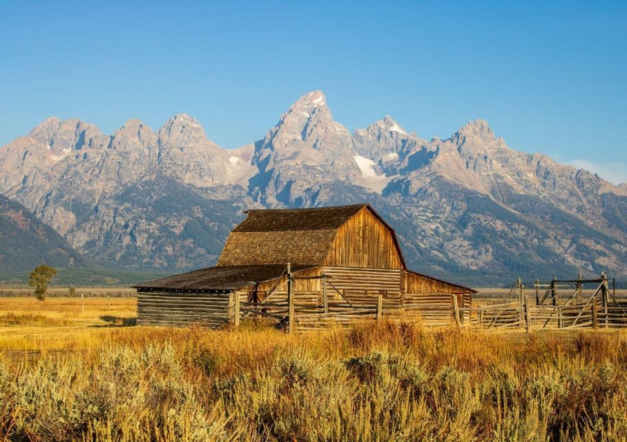 A rustic wooden barn stands in a golden field with a large, jagged mountain range in the background under a clear blue sky.