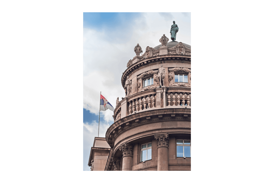 La bandiera serba sventola accanto alla cupola decorata di un edificio classico con statue, sotto un cielo parzialmente nuvoloso.