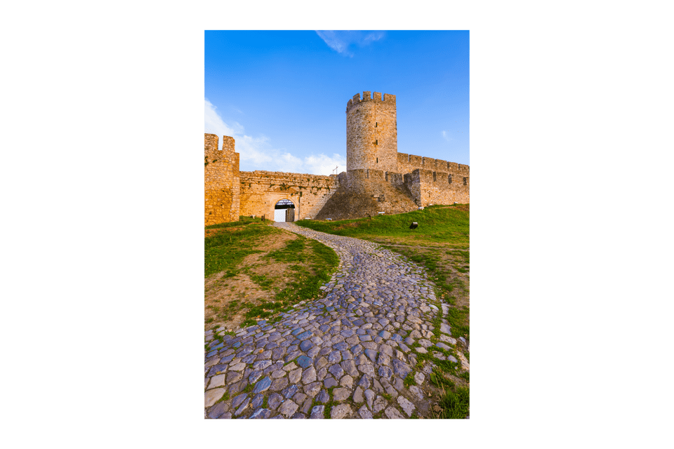 Un sentiero lastricato tortuoso sale su una collina erbosa fino al cancello di un'antica fortezza di pietra sotto un cielo blu.