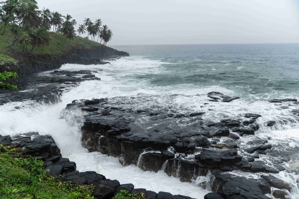 Le bianche onde dell'oceano si infrangono su una costa di roccia vulcanica nera, con una scogliera verde orlata di palme sullo sfondo, sotto un cielo nuvoloso.