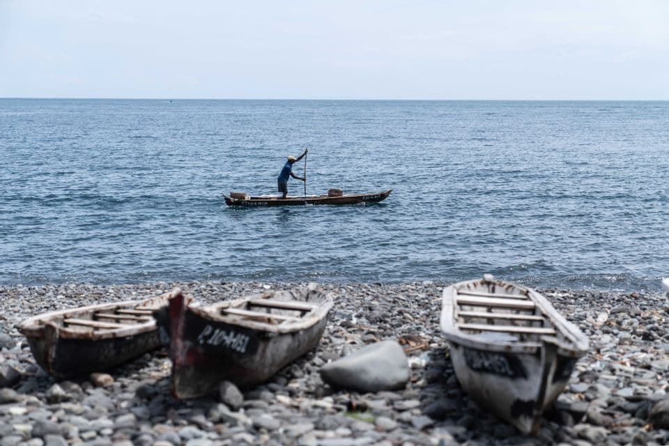 Un pescatore manovra una piccola imbarcazione sul mare, con tre vecchie barche di legno che riposano su una spiaggia rocciosa in primo piano.