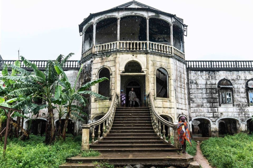 All'ingresso di una villa coloniale, logora, con una grande scalinata e piante tropicali rigogliose, si possono vedere persone.
