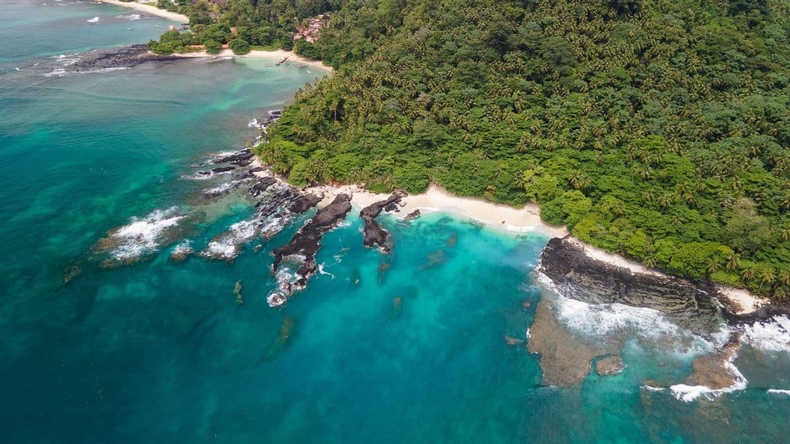 Una vista aerea di una costa tropicale dove una fitta giungla verde incontra l'oceano turchese, con rocce vulcaniche e una piccola spiaggia sabbiosa.